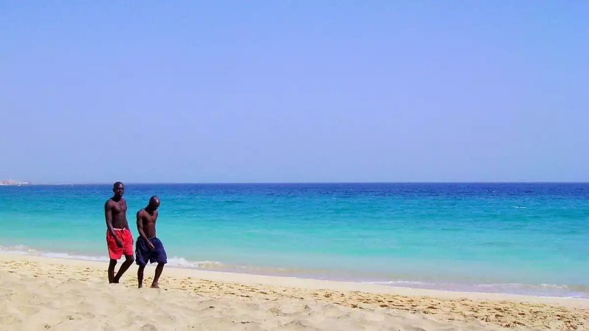 Due persone passeggiano lungo la spiaggia di Boa Vista, i loro passi lasciano dolci impronte sulla soffice sabbia, mentre il mare turchese e il cielo azzurro e limpido creano uno sfondo idilliaco.