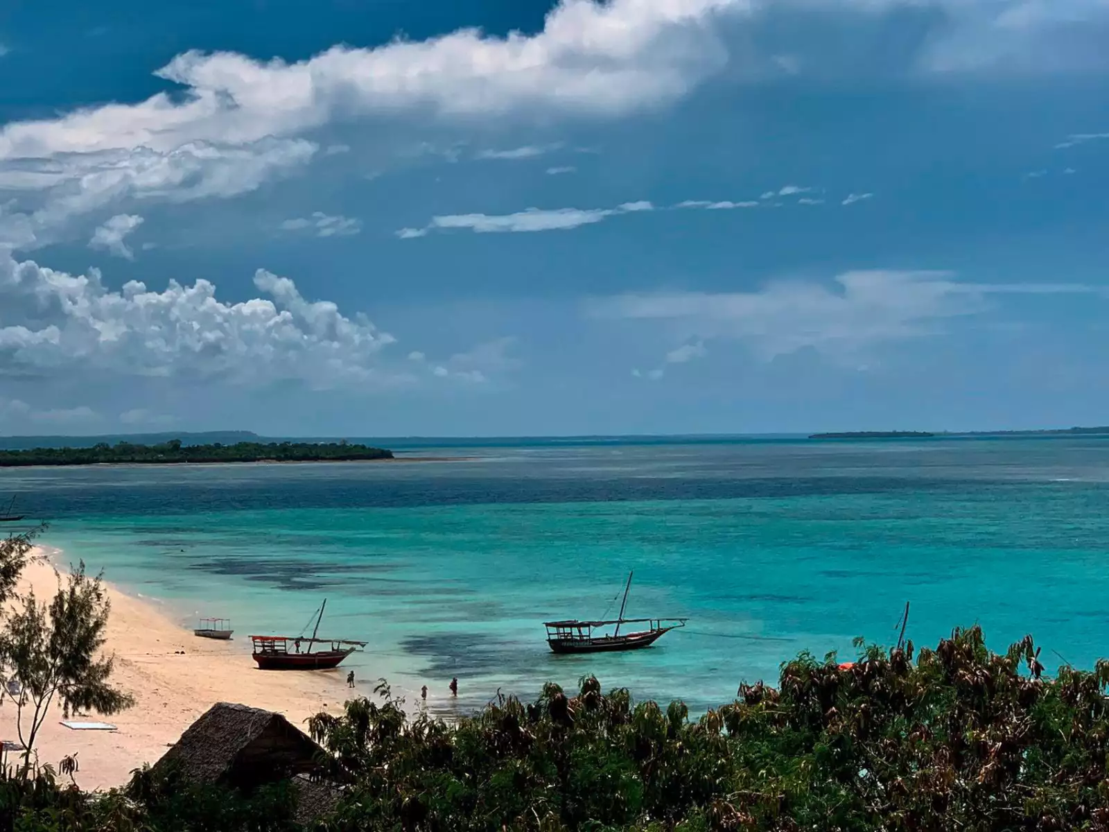 La scena della spiaggia tropicale al Waves Resort presenta acque turchesi e due piccole imbarcazioni adagiate sulla riva, circondate da una vegetazione lussureggiante. Un cielo nuvoloso incombe sullo sfondo, aggiungendo un tocco di drammaticità a questa idilliaca destinazione di Palumbo.