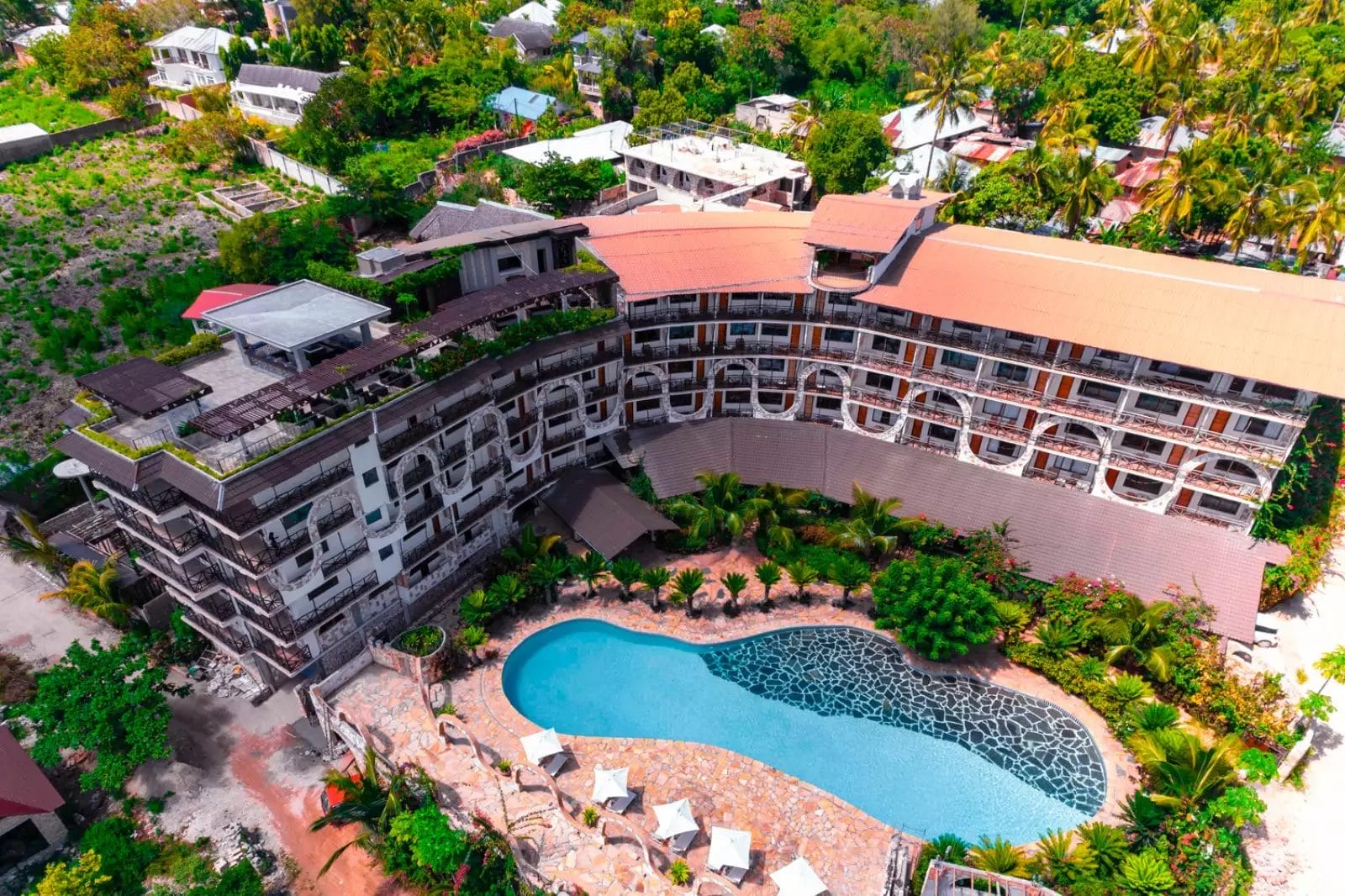 Vista aerea del Palumbo Waves Resort, un grande hotel dal design curvilineo, dotato di una piscina centrale all'aperto, circondata da sedie a sdraio e vegetazione lussureggiante in un ambiente tropicale.