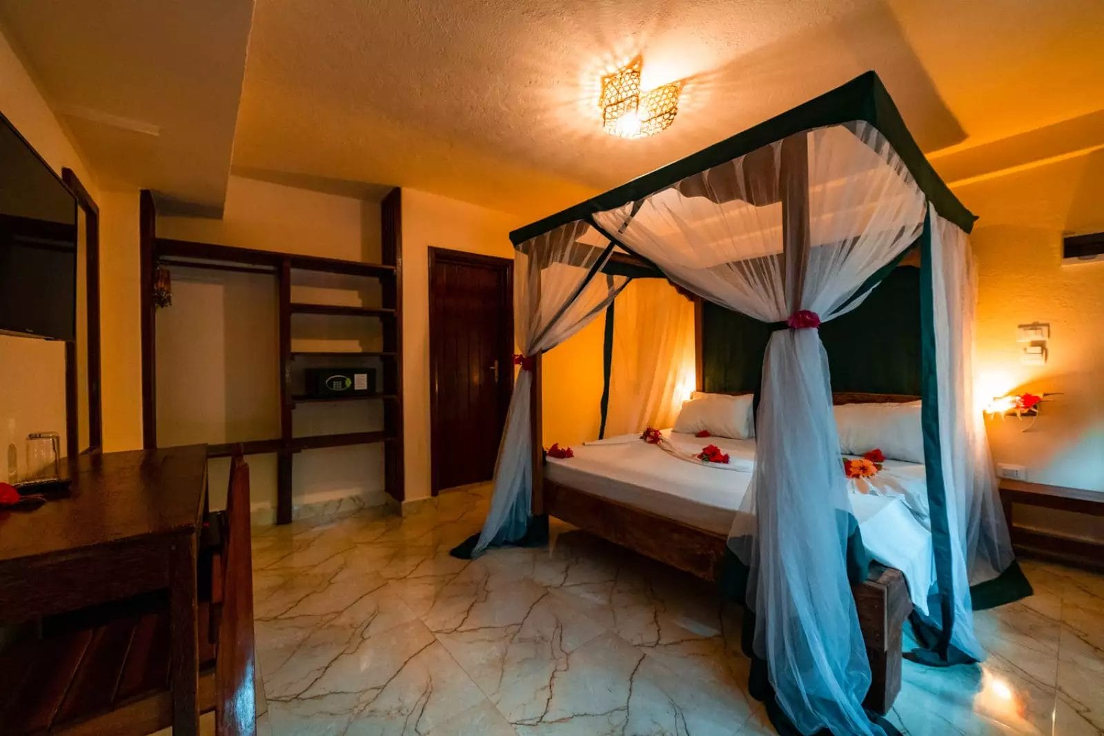 L'accogliente camera da letto del Palumbo Waves Resort è dotata di un letto a baldacchino avvolto da tende bianche, fiori tropicali che decorano il letto, mobili in legno e una calda luce soffusa.