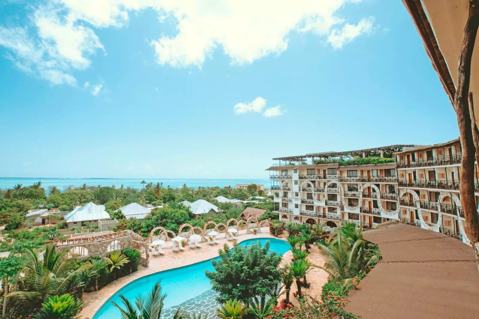 Vista aerea del Palumbo Waves Resort con una piscina scintillante, circondato da una vegetazione lussureggiante ed eleganti edifici, il tutto con vista sull'oceano infinito sotto un cielo azzurro e terso.