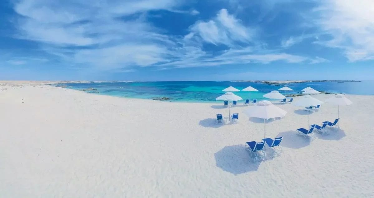 La spiaggia deserta di Alila Hinu Bay vanta sabbia bianca, sedie a sdraio blu e ombrelloni bianchi sotto un cielo parzialmente nuvoloso e un oceano turchese, offrendo un rifugio esclusivo.