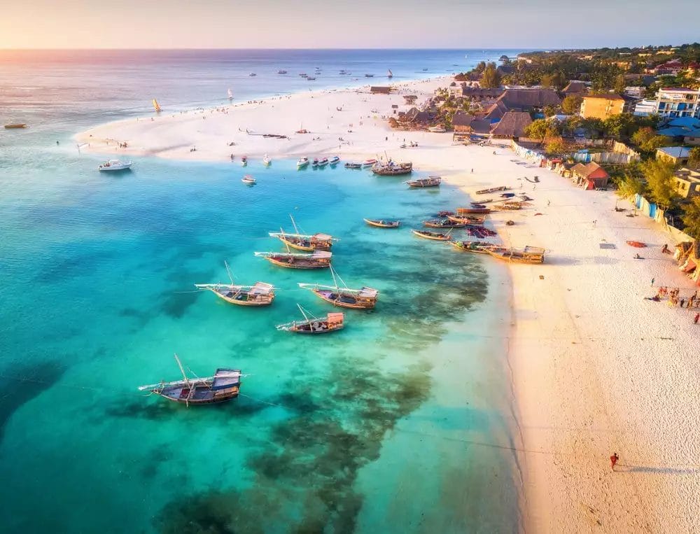 Vista aerea di una spiaggia al Palumbo Waves Resort con acqua turchese e limpida, diverse barche di legno vicino alla riva e persone che camminano lungo la spiaggia di sabbia bianca. Edifici e vegetazione lussureggiante abbelliscono lo sfondo.