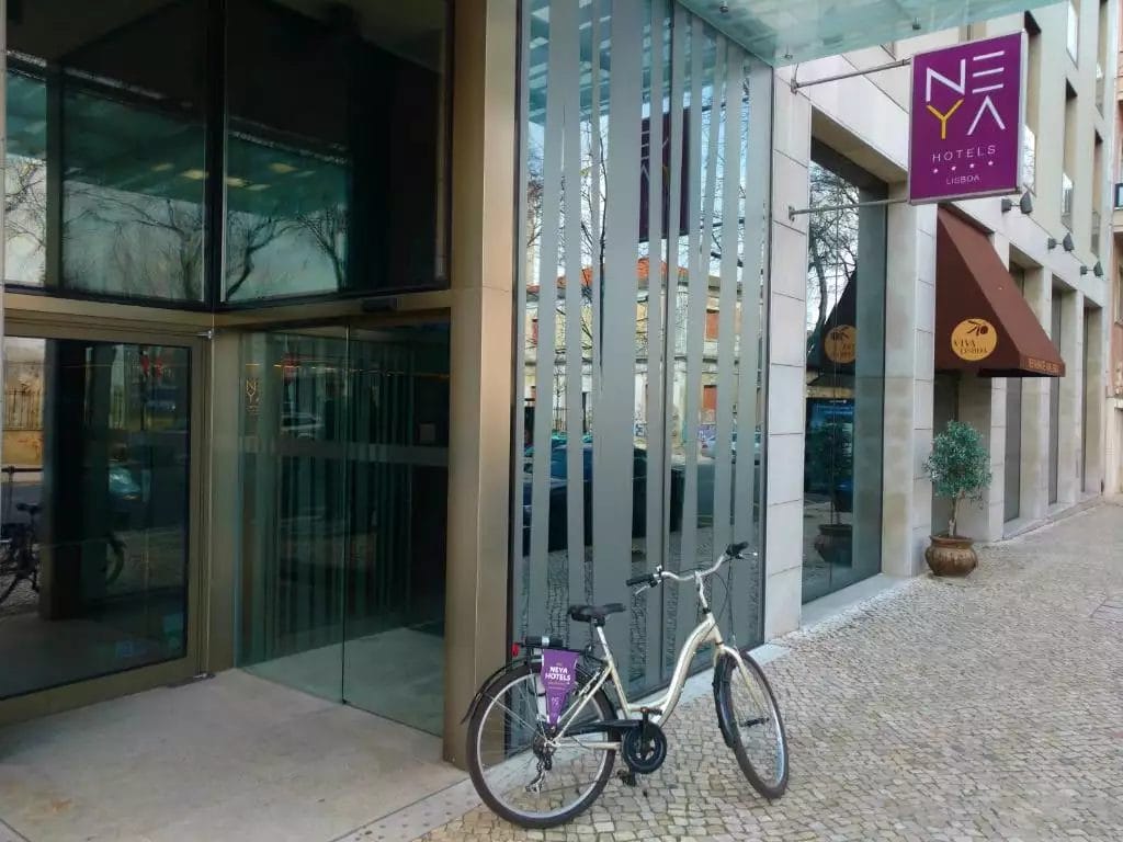 Una bicicletta è parcheggiata fuori dall'ingresso del Neya Lisboa Hotel, la cui facciata in vetro riflette la vibrante energia di Lisbona. La segnaletica del NEYA Hotels si erge orgogliosa sopra la porta, offrendo un caloroso invito a chi festeggia il Capodanno con stile.