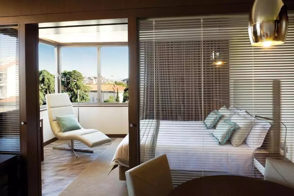 Questa camera da letto moderna, che ricorda lo stile chic del Neya Lisboa Hotel, è dotata di un grande letto e cuscini accanto a una comoda poltrona vicino alla finestra. Porte in vetro con persiane la separano dalla zona pranzo, consentendo a molta luce naturale di illuminare lo spazio.