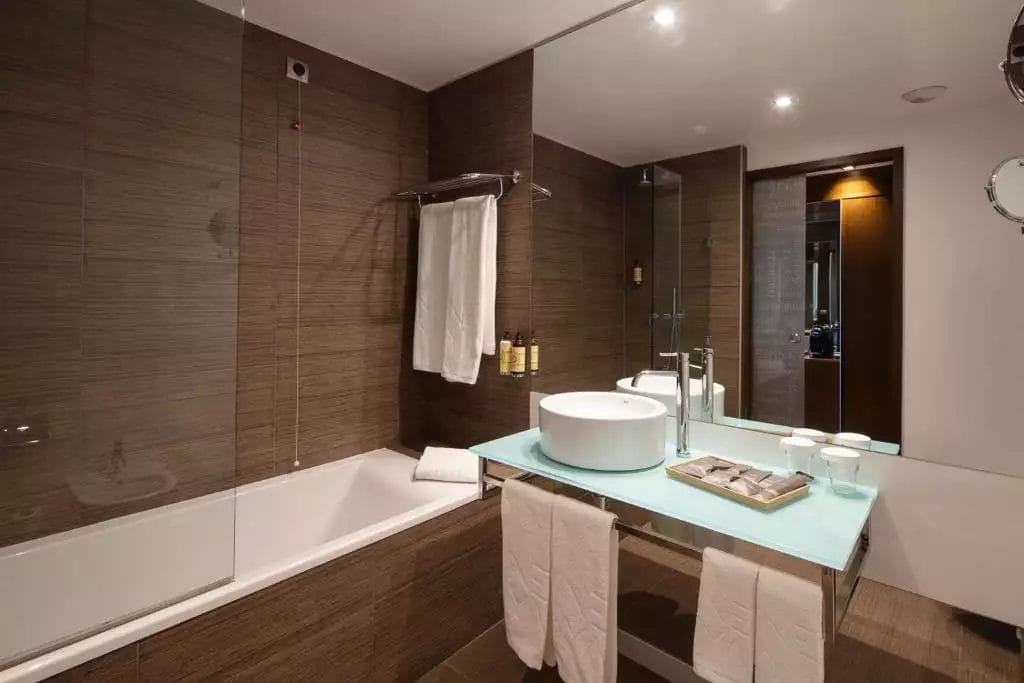 Bagno moderno che ricorda un hotel di lusso, con un grande specchio, una parete doccia in vetro, una vasca da bagno e un lavandino da appoggio. Gli asciugamani sono appesi a un'asta e sotto il lavandino. I prodotti da toeletta sono ordinatamente disposti sul piano di lavoro, invitandoti a rilassarti come se fossi a Lisbona durante i festeggiamenti di Capodanno.