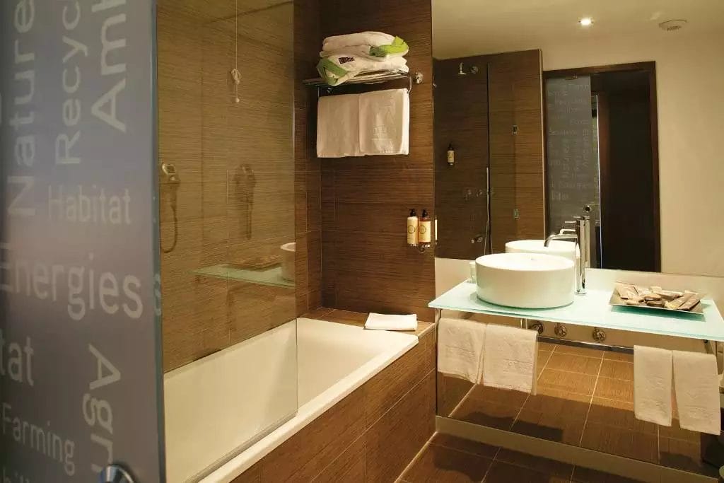 Bagno moderno presso l'hotel Neya Lisboa con dettagli in legno, vasca da bagno, doccia in vetro, lavandino rotondo su un piano in vetro e asciugamani ordinatamente impilati.