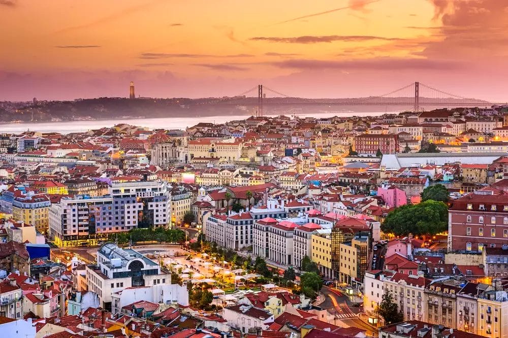 Vista aerea del vivace paesaggio urbano di Lisbona al tramonto, che mostra edifici colorati, strade trafficate e un grande ponte su un fiume. Perfetto per un'esperienza speciale di Capodanno, magari soggiornando al Neya Lisboa Hotel.