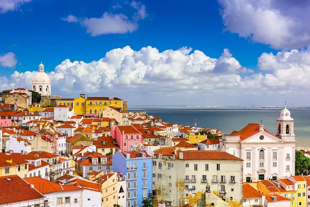 Vista di un colorato quartiere di Lisbona con edifici dai tetti rossi, una chiesa bianca e l'acqua in lontananza sotto un cielo parzialmente nuvoloso, che cattura il fascino vicino all'Hotel Neya Lisboa.