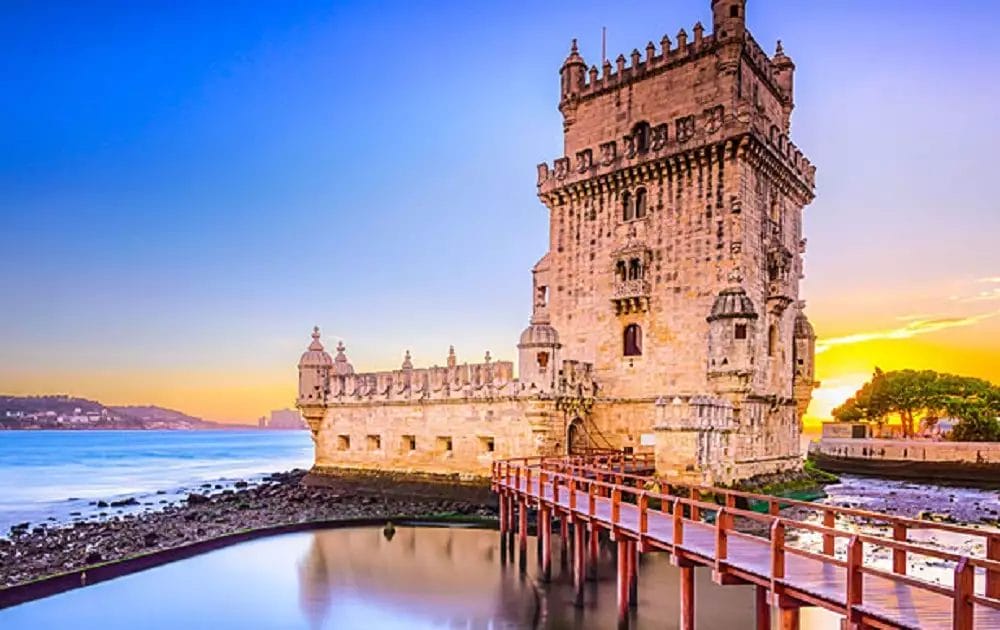 L'immagine cattura la Torre di Belém a Lisbona, Portogallo, al tramonto. Questa storica struttura in pietra sul fiume Tago è splendidamente illuminata, con una passerella in legno che conduce al suo ingresso. Nelle vicinanze, il Neya Lisboa Hotel offre una vista perfetta per chi festeggia Capodanno con stile.
