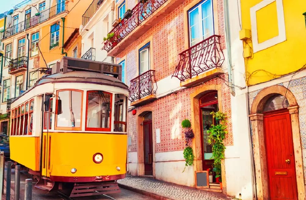 Un tram giallo scivola lungo una strada acciottolata, il cui percorso si snoda tra edifici colorati e balconi decorati in uno scenario così pittoresco che potrebbe essere scambiato per una bozza automatica.