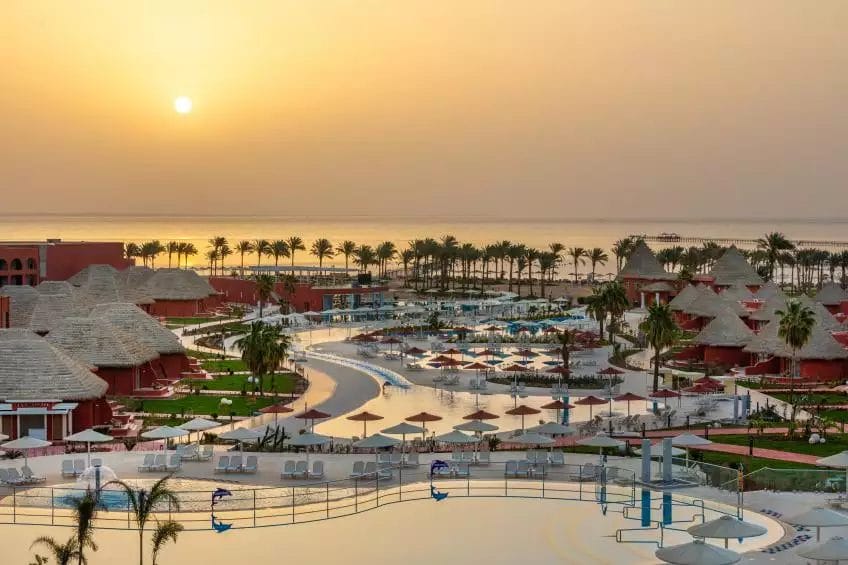 Un resort in riva al mare al tramonto, con palme, ombrelloni e piscine all'aperto sotto un cielo dorato, offre una fuga serena nel paradiso dell'ALBATROS.