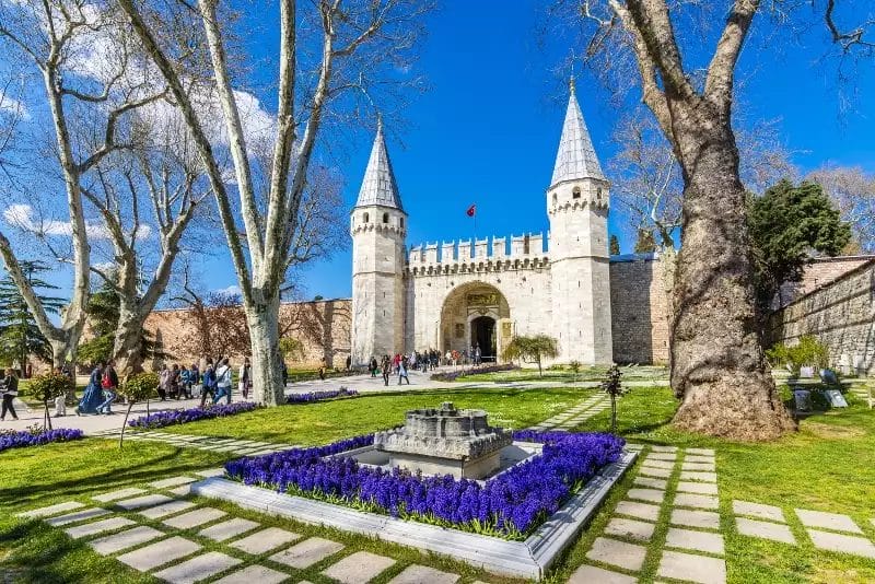 Storico edificio in pietra con torri coniche e una porta centrale, che ricorda il fascino della Cappadocia, circondato da un giardino paesaggistico con fiori viola e una fontana sotto il cielo azzurro di Istanbul. La gente passeggia lì vicino, magari godendosi un mini tour dell'incantevole scenario.