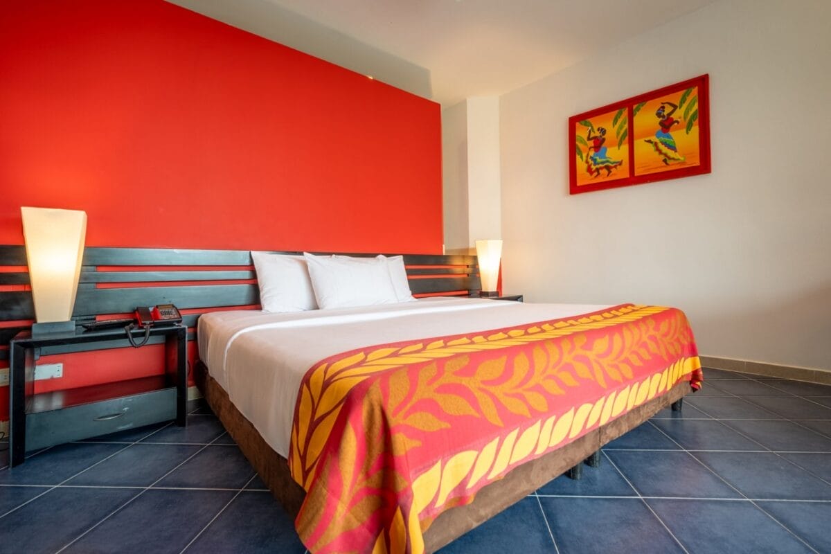 Camera d'albergo al Bravo Decameron Baru con un letto grande, una parete con accenti rossi, una coperta colorata e un vivace dipinto a forma di pappagallo sopra il letto.