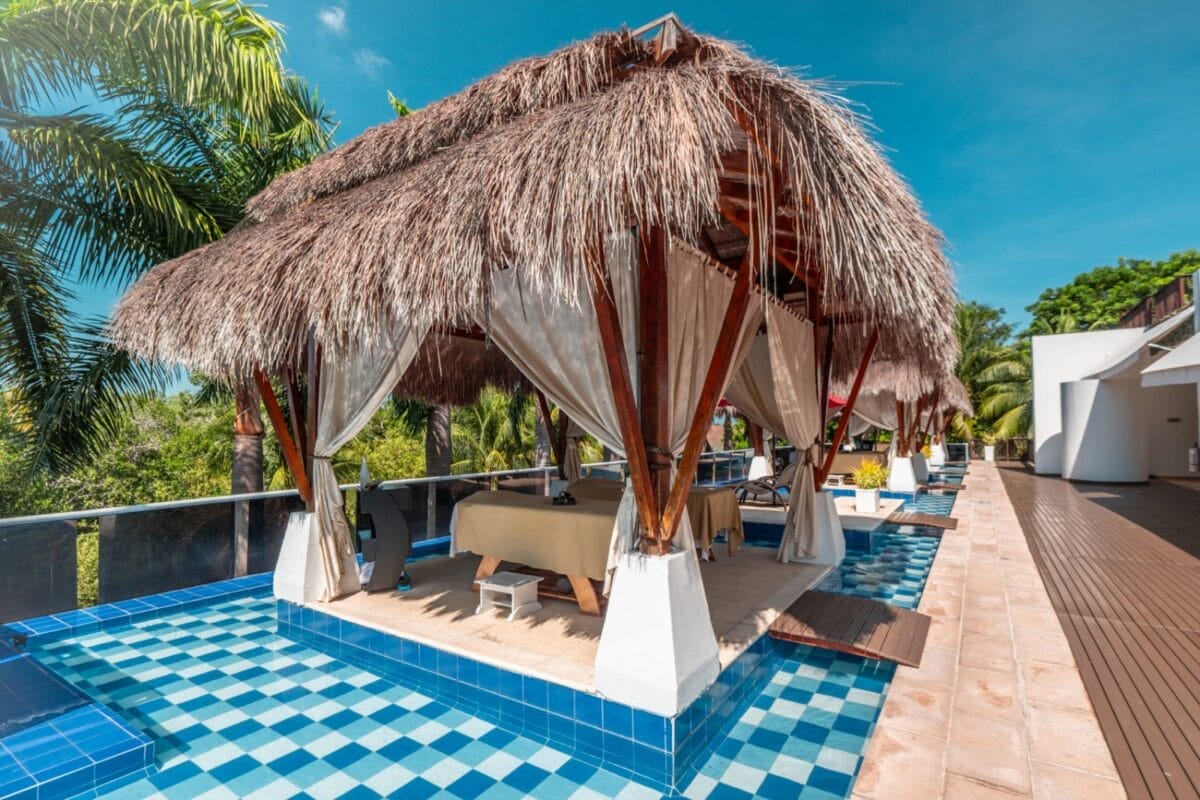 Una cabana con tetto di paglia e tende si trova sopra una piscina rivestita di piastrelle blu circondata da palme al BRAVO DECAMERON BARU.