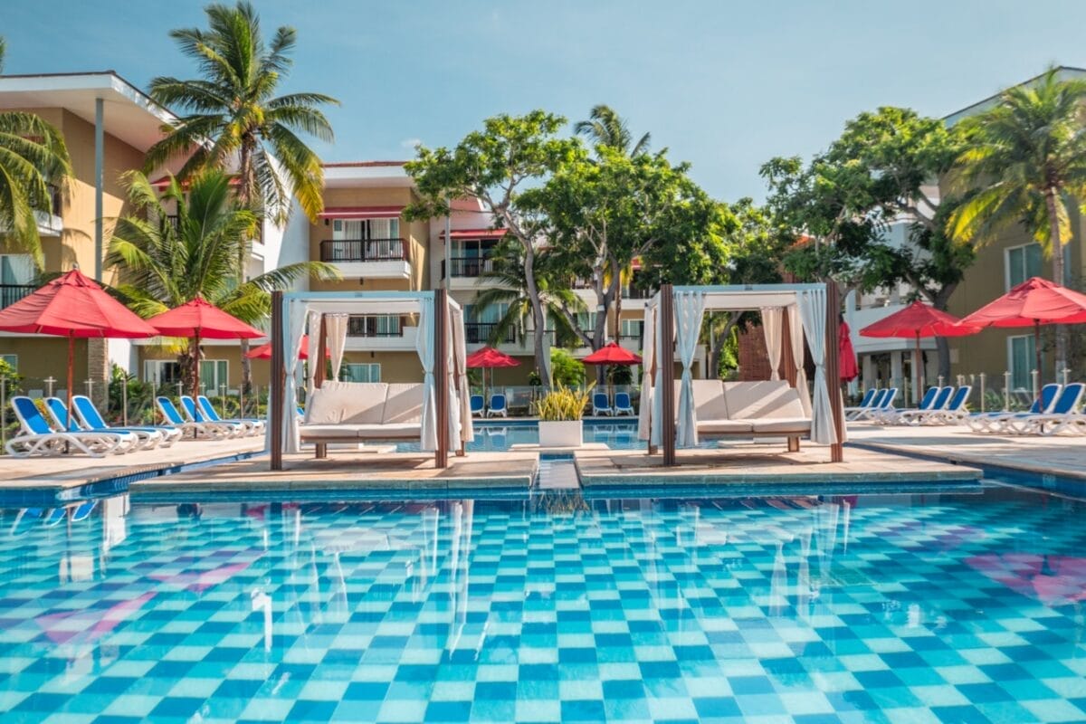 Le cabine a bordo piscina con lettini, ombrelloni rossi e palme creano un'oasi di relax al BRAVO DECAMERON BARU, caratterizzato da piastrelle a scacchi blu che creano una vivace atmosfera da resort.