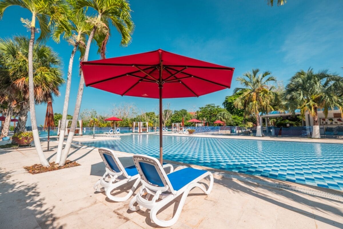 Due sedie a sdraio sotto un ombrellone rosso accanto alla piscina piastrellata di blu e bianco del BRAVO DECAMERON BARU, circondata da palme e cielo terso.