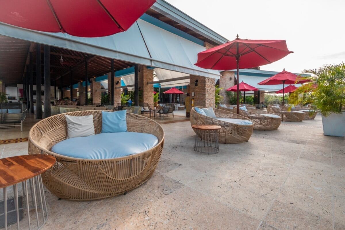 Patio esterno al BRAVO DECAMERON BARU con sedie a sdraio in vimini, cuscini blu, ombrelloni rossi e tavolini laterali su un elegante pavimento in pietra.