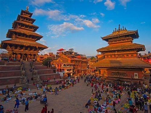 Una piazza affollata in Nepal con antichi templi a pagoda e persone che fanno acquisti nei mercati all'aperto sotto un cielo azzurro.
