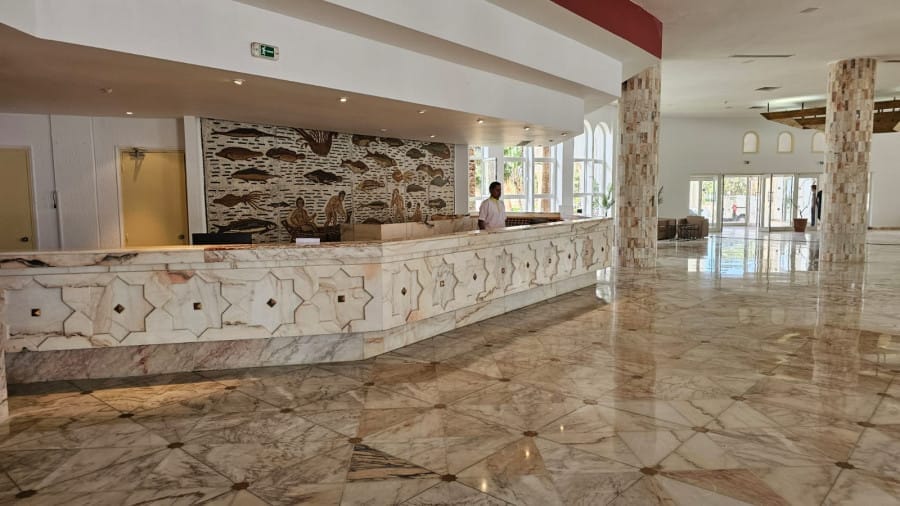 Ampia hall dell'hotel con pavimenti in marmo e un lungo bancone della reception presso il NICOLAUS CLUB EL FATIMI a Mahdia; una persona in piedi dietro il bancone accoglie calorosamente gli ospiti vicino all'Acqua Park Mahdia.