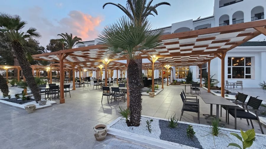 Area salotto all'aperto del ristorante con pergole in legno, palme, tavoli e sedie vicino a un edificio bianco al tramonto presso il NICOLAUS CLUB EL FATIMI a Mahdia.