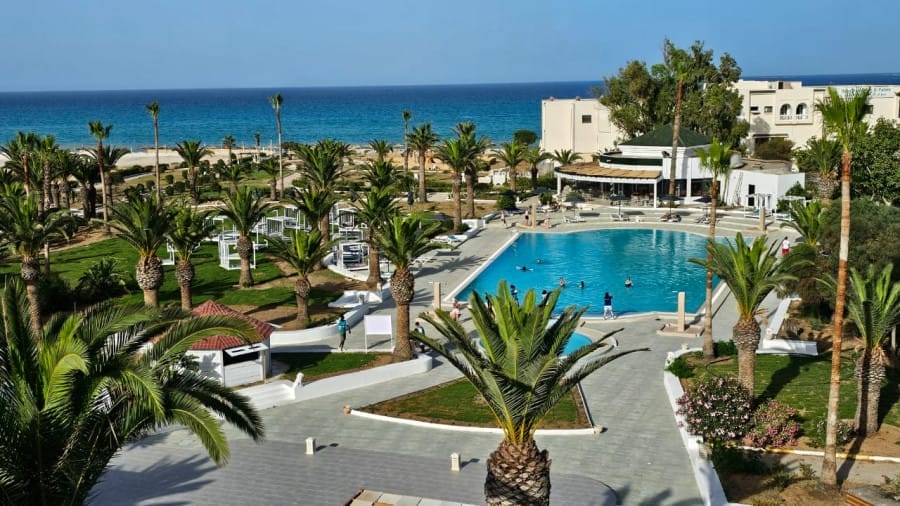 L'area della piscina del resort NICOLAUS CLUB EL FATIMI, circondata da palme e frequentata da persone che nuotano, si affaccia sul mare e sul cielo azzurro e limpido a Mahdia, vicino all'ACQUA PARK MAHDIA.