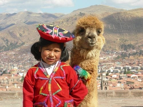 Una ragazza in abiti tradizionali andini è in piedi accanto a un lama, con una città peruviana e le montagne sullo sfondo: uno scenario invitante per il tuo tour esclusivo in PERÙ 2025.