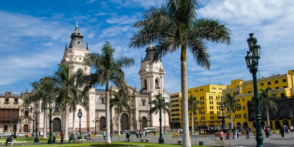 Palme e persone in una piazza con una grande cattedrale ed edifici gialli sotto un cielo azzurro: scopri il fascino del Perù in un tour indimenticabile nel 2025.