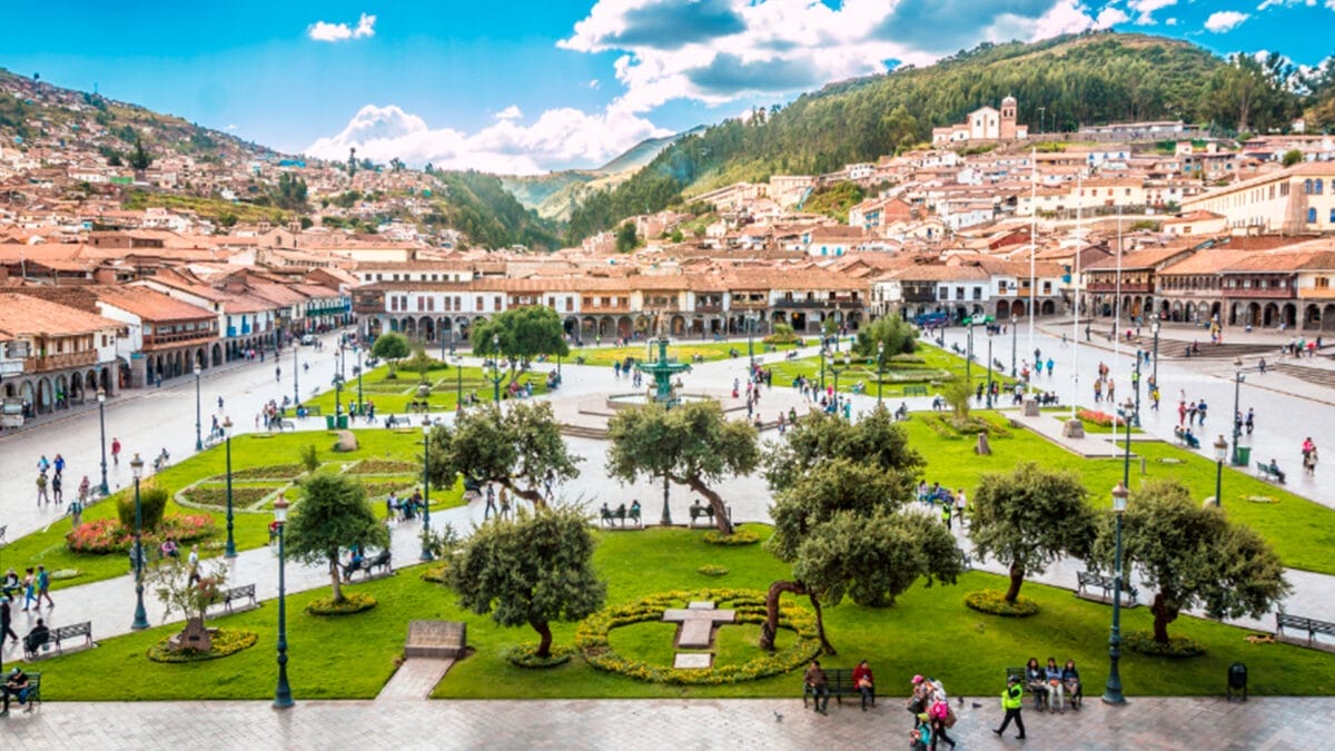 Una piazza cittadina in Perù con prati verdi, alberi e persone che camminano, sullo sfondo delle montagne sotto un cielo azzurro: un luogo perfetto per godersi un tour esclusivo nel 2025.