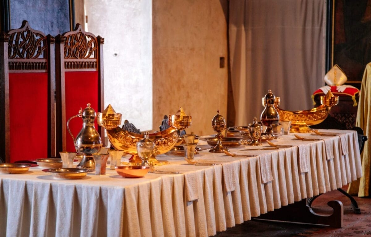 Un lungo tavolo con stoviglie dorate e sedie con cuscini rossi evoca l'eleganza degli storici CASTELLI DELLA BOEMIA, perfetti per una raffinata esperienza di PRAGA TOUR.