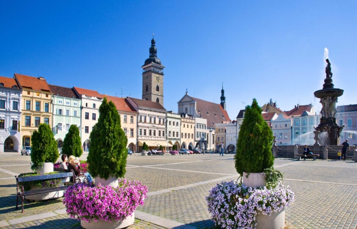 Piazza della città con edifici colorati, un'alta torre dell'orologio, una fontana e fioriere sotto un cielo azzurro e limpido: l'ideale per un tour di Praga alla scoperta degli affascinanti Castelli della Boemia.