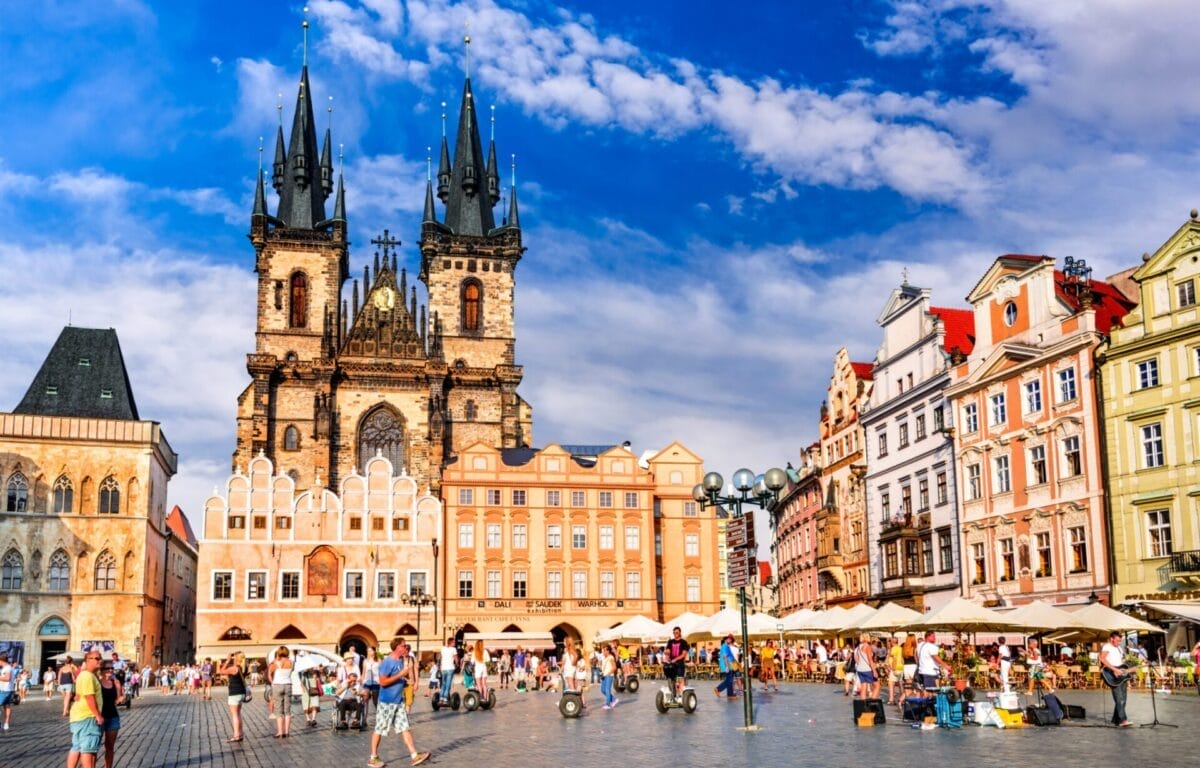 Persone che passeggiano in una vivace piazza con edifici colorati e una chiesa gotica sotto un cielo azzurro a Praga, perfetta per un tour alla scoperta dei Castelli della Boemia.