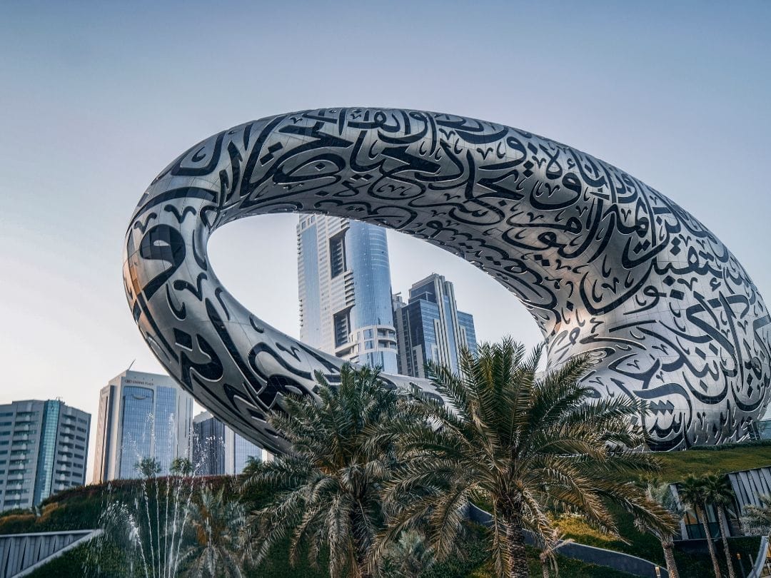 Edificio ovale argentato con calligrafia araba, circondato da palme e grattacieli della città: scoprite questo iconico punto di riferimento durante un'avventura TOUR DUBAI EXPRESS.