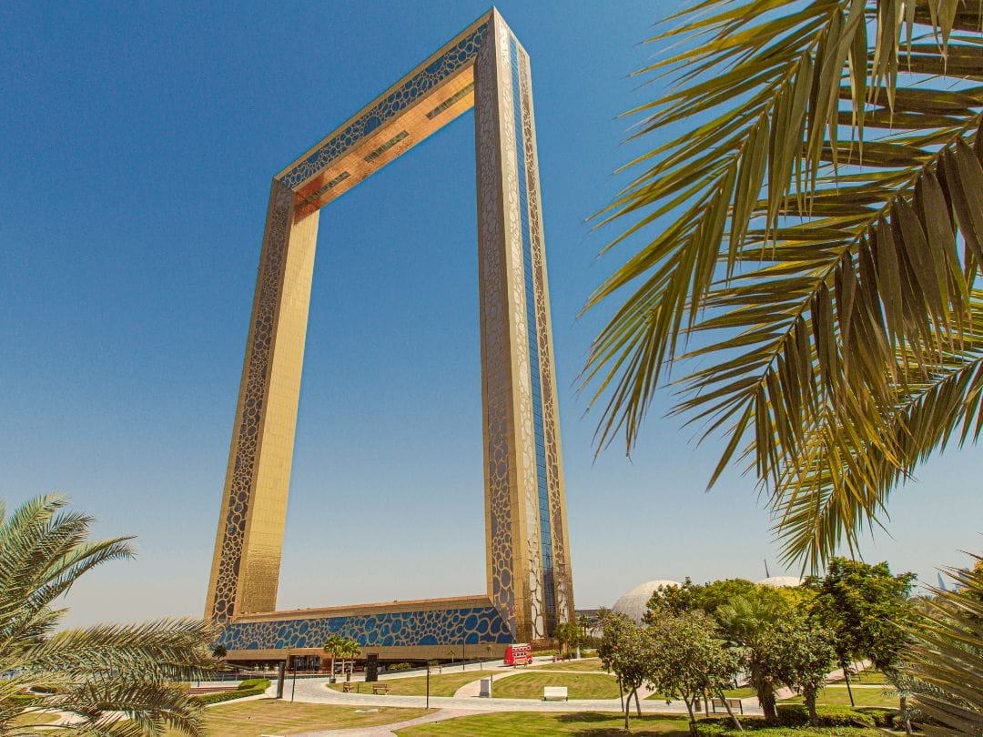 Una grande struttura rettangolare dorata si erge tra le palme e il verde, sotto un cielo azzurro e limpido: un'attrazione imperdibile in ogni avventura del TOUR DUBAI EXPRESS.