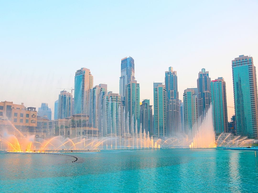 Alti grattacieli circondano una grande fontana blu con getti d'acqua che spruzzano in aria al tramonto, creando una vista mozzafiato perfetta per la vostra avventura TOUR DUBAI EXPRESS.