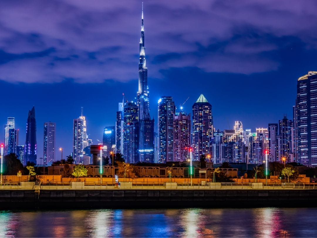 Lo skyline di Dubai di notte con i grattacieli illuminati che si riflettono nell'acqua sotto un cielo viola: vivete tutto questo in un'avventura TOUR DUBAI EXPRESS.
