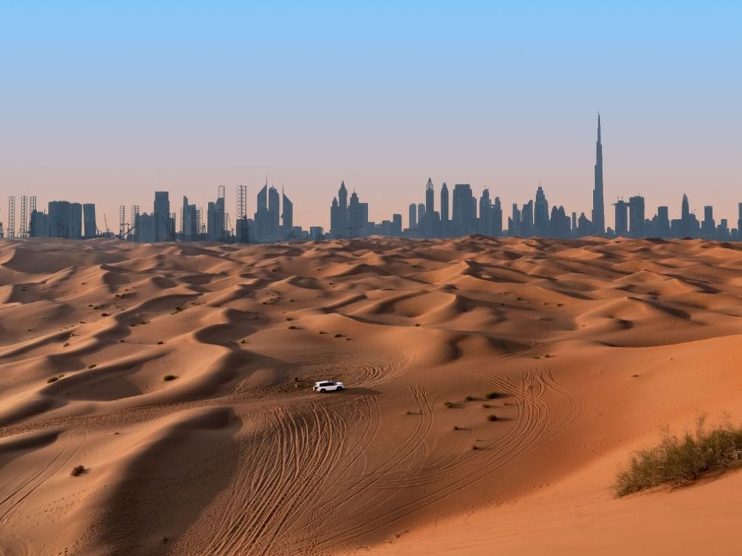 Un SUV bianco attraversa le dune del deserto durante un tour di Dubai, con l'iconico skyline della città sullo sfondo sotto un cielo limpido.