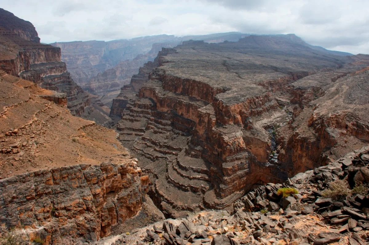 Canyon rocciosi stratificati si estendono in lontananza sotto un cielo nuvoloso, con scogliere frastagliate e valli profonde.