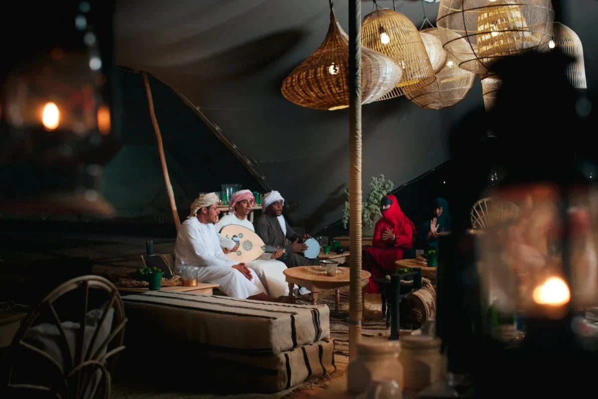 Un gruppo di persone in abiti tradizionali si riunisce all'interno, uno suona un oud, con calde lanterne appese sopra.