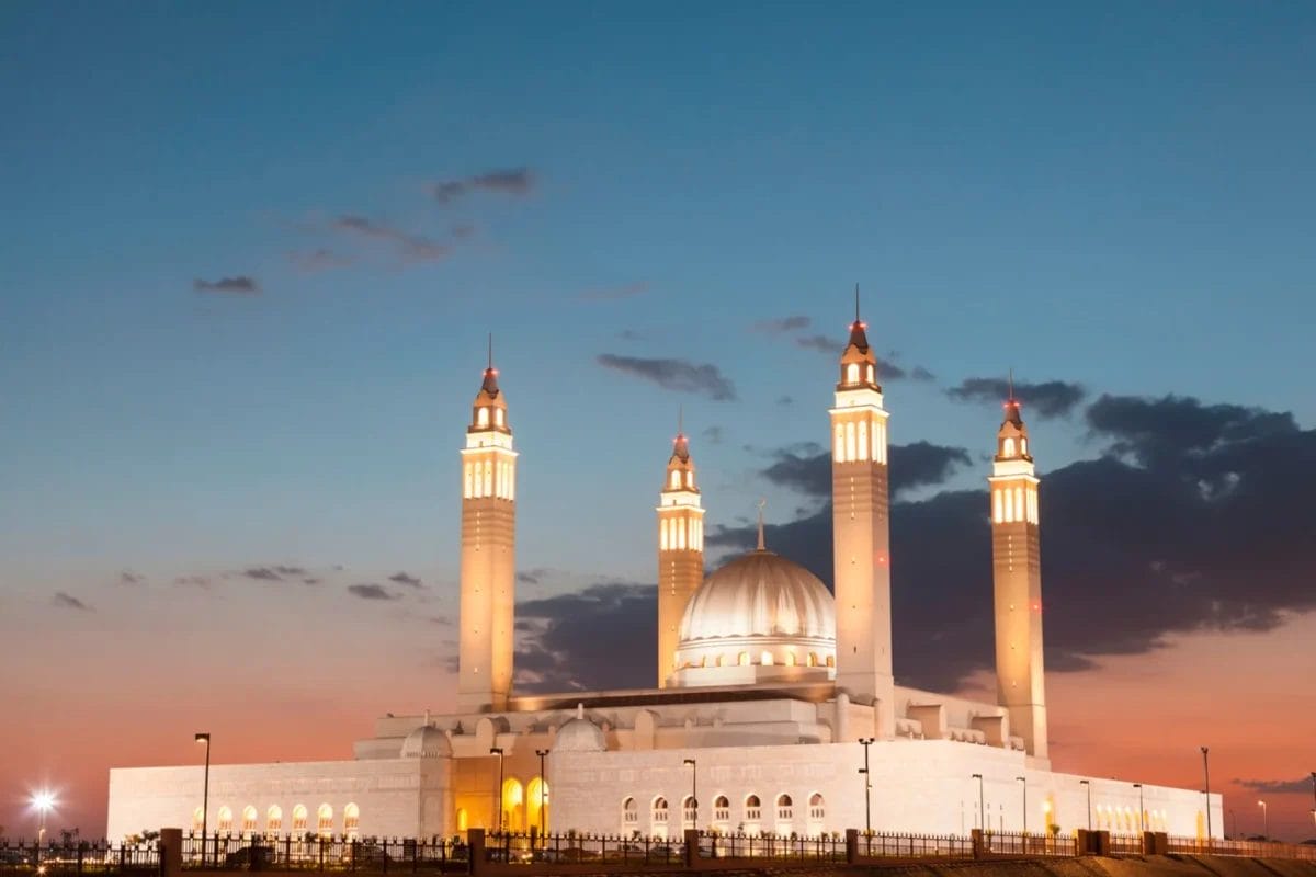 Una grande moschea bianca con quattro minareti e una cupola, illuminata al tramonto contro un cielo colorato.
