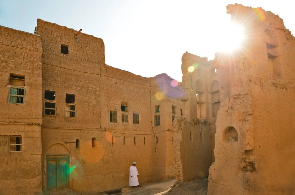 Una persona vestita di bianco si trova vicino a vecchi edifici di adobe illuminati dal sole, con il sole che filtra da un muro in rovina.