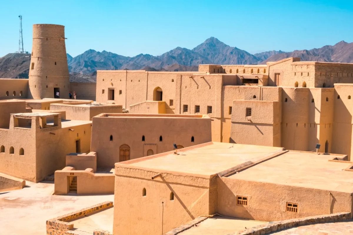 Antica fortezza abbronzata con torri e montagne sullo sfondo, sotto un cielo azzurro e limpido.