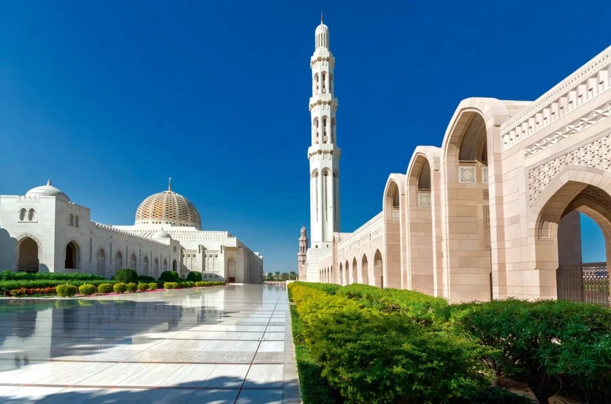 Una grande moschea con una grande cupola, un alto minareto, corridoi ad arco e una vegetazione lussureggiante sotto un cielo azzurro e limpido.