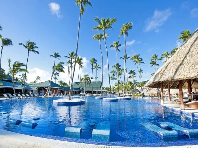 Grande piscina all'aperto con sedie a sdraio, palme e un bar con tetto di paglia sotto un cielo azzurro e limpido al Barcelo Bavaro Beach.