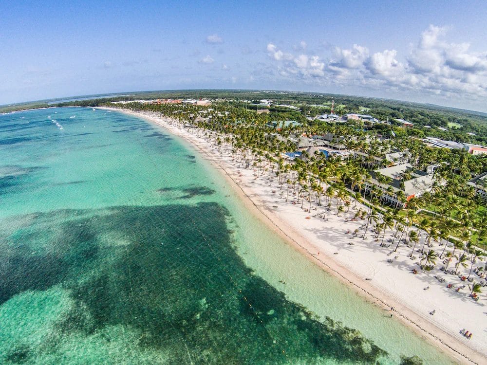 Vista aerea della SPIAGGIA DI BARCELO' BAVARO, con acqua turchese, sabbia bianca e palme sotto un cielo parzialmente nuvoloso.