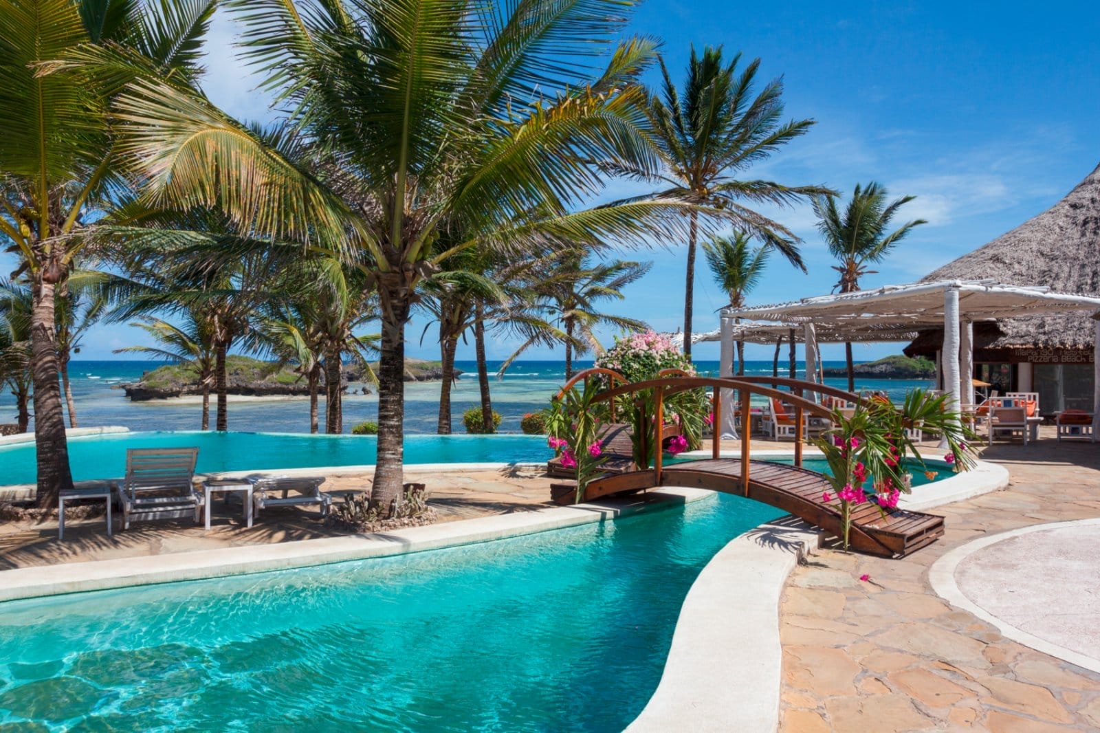 Piscina tropicale del Lily Palm Resort con palme, un piccolo ponte di legno e vista sull'oceano sotto un cielo azzurro e limpido.