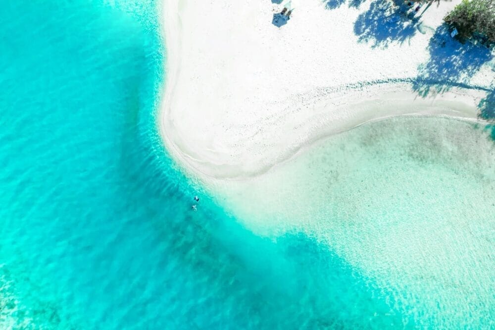 Veduta aerea della spiaggia di sabbia bianca dello SKY BEACH HOTEL che incontra un'acqua turchese e trasparente, con onde delicate. Le ombre degli alberi si proiettano sulla sabbia mentre due persone nuotano vicino alla riva.