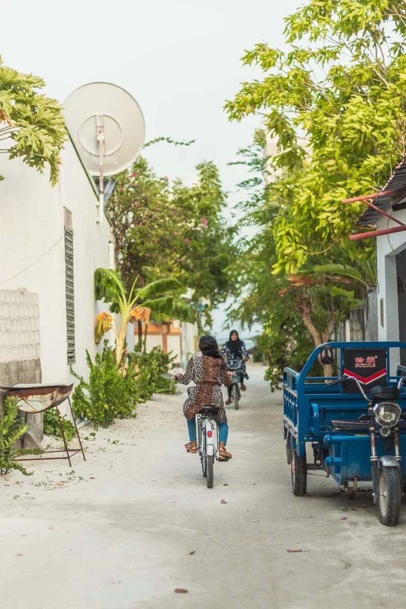 Due persone vanno in bicicletta lungo una strada stretta e alberata con muri bianchi su entrambi i lati. Un veicolo blu a tre ruote è parcheggiato sulla destra e lungo la strada, vicino allo SKY BEACH HOTEL, sono visibili il verde e le antenne paraboliche.