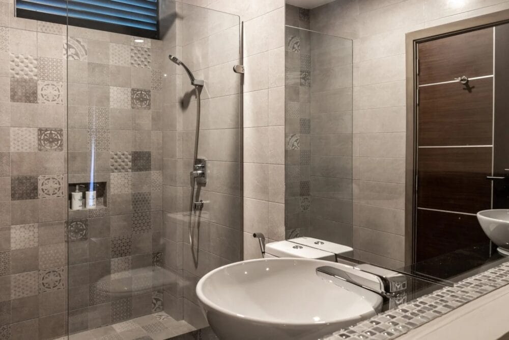 Bagno moderno dello SKY BEACH HOTEL con doccia chiusa da un vetro, piastrelle grigie a fantasia, lavandino rotondo bianco su un piano d'appoggio scuro e porta in legno; la luce naturale entra da una piccola finestra.