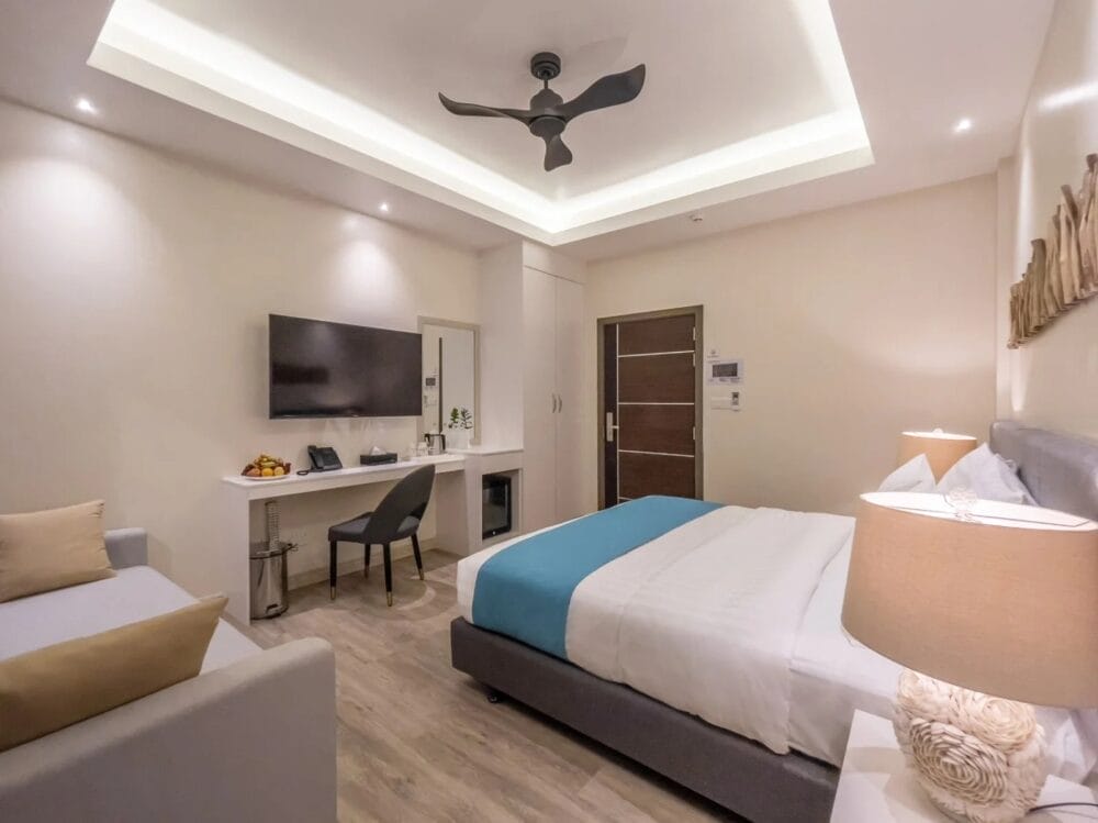 Moderna camera d'albergo dello SKY BEACH HOTEL con un letto ben fatto in lenzuola bianche e blu, un divano, una grande TV a parete, una scrivania e una sedia, un ventilatore a soffitto e un'illuminazione calda. Lo spazio è caratterizzato da un design pulito e minimalista.